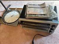 Photo showing the Hamilton Beach toaster oven alongside the Bulova wall clock, the toaster oven has the manual and packaged accessories on top, and both items are placed on a carpeted floor.