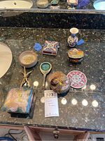 Photo showing eight assorted trinket boxes and small decorative items displayed on dark marble countertop. Items include cloisonné boxes, vases, and a decorative matchbox holder with tongs.