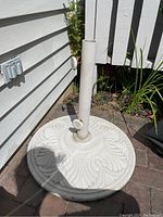 Full view of a white decorative outdoor umbrella stand with a round embossed leaf pattern base, placed outdoors near a house.