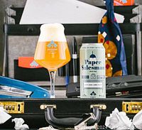 Dominion City branded tulip glass filled with beer and a can of Paper Salesman Pale Ale displayed on a black case alongside office supplies and tie.