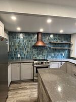 Example of blue hand-glazed rectangular ceramic tile backsplash installed behind a stainless steel range and copper hood, with gray quartz countertops and white shaker cabinets.