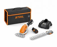 STIHL HSA 26 shrub shears kit showing handheld tool with shrub blade attached, grass trimming blade, battery, charger, and carrying case.