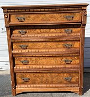 Front view of the antique five drawer chest showing walnut wood with burl paneling on drawer fronts, brass bail handles, and top carved molding detail.