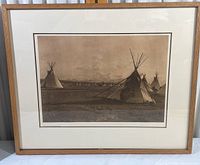 Framed sepia-tone print showing several Native American tipis in an open field under cloudy sky, inside wooden frame with matting.
