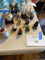 Overview of 12 mini Asian inspired porcelain jugs and vases with varying colors and designs, arranged on a table with some on wooden stands and one metal pagoda figurine.