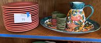 Stack of 14 salmon colored dinner plates, hand-painted pitcher, matching serving tray, small glasses, and serving dish on a shelf