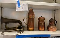 Full view of all 5 items on shelf showing 3 copper pieces: pitcher, container, teapot; 2 brass horns stacked beside them.