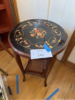 Photo showing round lacquered wood end table with a floral and scrollwork painted top surface, seating on wooden floor.