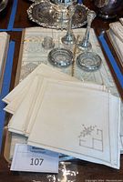 Stack of 18 embroidered linen napkins on top of a European linen tablecloth with silver and crystal tableware in background.