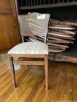 Photo of one upright wooden folding chair with cane back and upholstered floral patterned seat in front of a stack of similar chairs.