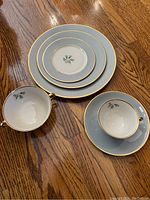 Photo showing samples of the plates in different sizes alongside matching tea cups and saucers with floral pattern and gold trim, set on wooden floor.