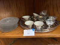 Silver serving tray with silver holder coffee mugs and etched glass cups on the tray, stacked glass plates beside it on wooden shelf