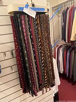 Photo showing a selection of men's ties hanging on a display rack, featuring various patterns including geometric and floral in burgundy, navy, and brown tones.