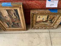 Two framed oil paintings displayed side by side next to a red patterned textile background, showing size tags and frames.