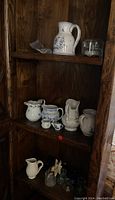 Display of assorted small porcelain pitchers and bud vases on wooden shelving, with a cut crystal music box visible