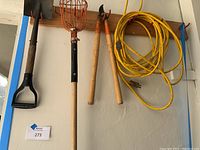 Small shovel, orange fruit picker, bolt cutters, and yellow extension cord hung on a hook rack on wall. All items for yard use, showing moderate use.