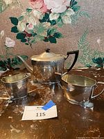 Silver plated teapot, sugar bowl, and creamer displayed on a marble surface with a floral wallpaper background.