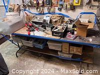 Wide view of workbench with tools and boxes scattered on top and underneath including clamps and containers of screws and nails.