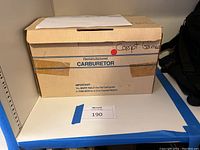 Cardboard box labeled “Remanufactured Carburetor” with handwritten “Comp. Games” on side, on shelf marked 190
