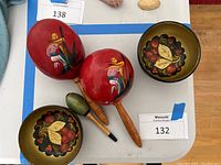 Two red hand painted wooden maracas with floral and bird designs, alongside two wooden bowls painted with floral and leaf motifs.