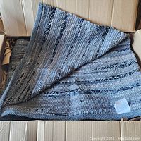 Photo showing the folded area rug inside a cardboard box, highlighting the pattern and texture of the blue and gray handwoven rag material.