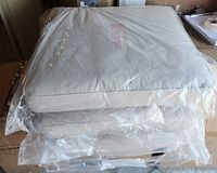 Three new patio sofa cushions, gray, wrapped in plastic stacked on each other.