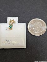 Photo of a small green gemstone pendant set in 14K yellow gold, displayed on a white mount with '14K GOLD' label, next to a dime coin for size comparison.