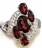 Close-up photo of a sterling silver ring featuring a central oval garnet, surrounded by four pear-shaped garnets and clear accent stones on the band. Shows details of the gemstones and silverwork.