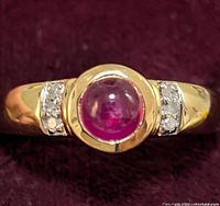 Close-up image of sterling silver ring set with round cabochon ruby center and white topaz side stones on a burgundy background.