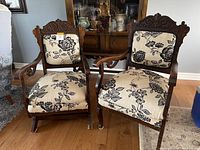 Pair of antique wooden chairs featuring beige fabric cushions with black floral pattern, placed on wooden floor in front of wood cabinet.