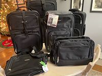 Five black luggage pieces arranged on and around a white table in living room. Three have wheels; two are smaller shoulder or duffel bags.
