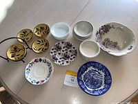 Photo showing assortment of small ceramic dishes on a table, including gold-colored leaf decorated plates, several white bowls, and decorative floral and scenic plates, plus a large scalloped bowl.