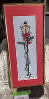 Full view of cross stitched Christmas lamp post wall hanging with gold frame and red matting, showing the decorative lamp post with wreath and snow base.