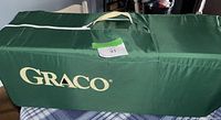 Graco branded green fabric carrying case for playpen with handle and zipper closure, placed on a plaid tablecloth.