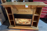 Full view of the large electric fireplace showing oak finish, open shelves on both sides, decorative carving above the firebox, and ember glow logs inside.