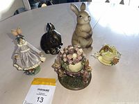 Overview of five rabbit figurines on a table: standing brown rabbit, glossy dark brown rabbit, rabbit in purple dress, floral centerpiece with rabbits, and yellow teacup with rabbit decor.