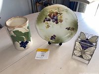 Three items on table including pottery vase with grapes, decorative plate with grapes, and butterfly glass candle holder.