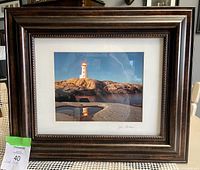 Front view of the framed signed photography print showing a white lighthouse with a red light on top, reflected in water on rocky terrain under a clear blue sky.