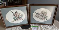 Both framed vintage cross stitch artworks placed side by side on a table.