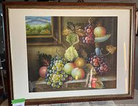 Framed wall art print showing a still life arrangement of grapes, apples, and other fruits with a small window landscape in the background. Visible signature on the print.