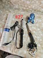 Photo showing rolled white tub mat sealed in packaging, black Conair curling iron, metallic curling iron, and small blue handheld massager, all placed on a beige polka dot surface.