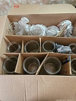 Cardboard box with compartments holding 12 clear glass mason jars of various sizes, some wrapped in paper for protection.