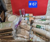 Photo showing multiple stacks and packages of disposable wooden forks, knives, spoons, paper cups with green labels, paper plates, napkins, and rolls of paper straws.