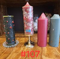 Four assorted pillar candles on wooden surface: a detailed red carved candle on brass holder, a black candle with red berries on glass base, a plain pink candle, and a plain blue-gray candle