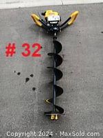 Full length image of gas powered ice auger showing black auger bit with cutting blade and yellow handles, taken on concrete surface.