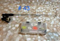 Ice fishing rod with small spinning reel alongside plastic tackle box of assorted lures and soft plastic baits.