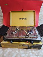 Martin propane camp stove with two burners open, showing stainless steel surface, burners, control knobs, and side wind guards.