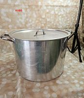 Photo of large aluminum stock pot with a lid and side handles against a beige bokeh background.