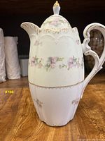 Side view of tall porcelain teapot showing floral pattern, scalloped lid, ornate handle with gold accent, and overall shape.