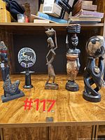 Five African themed small figurines displayed on wooden surface and shelf, showing variety including carved wood and beaded work.